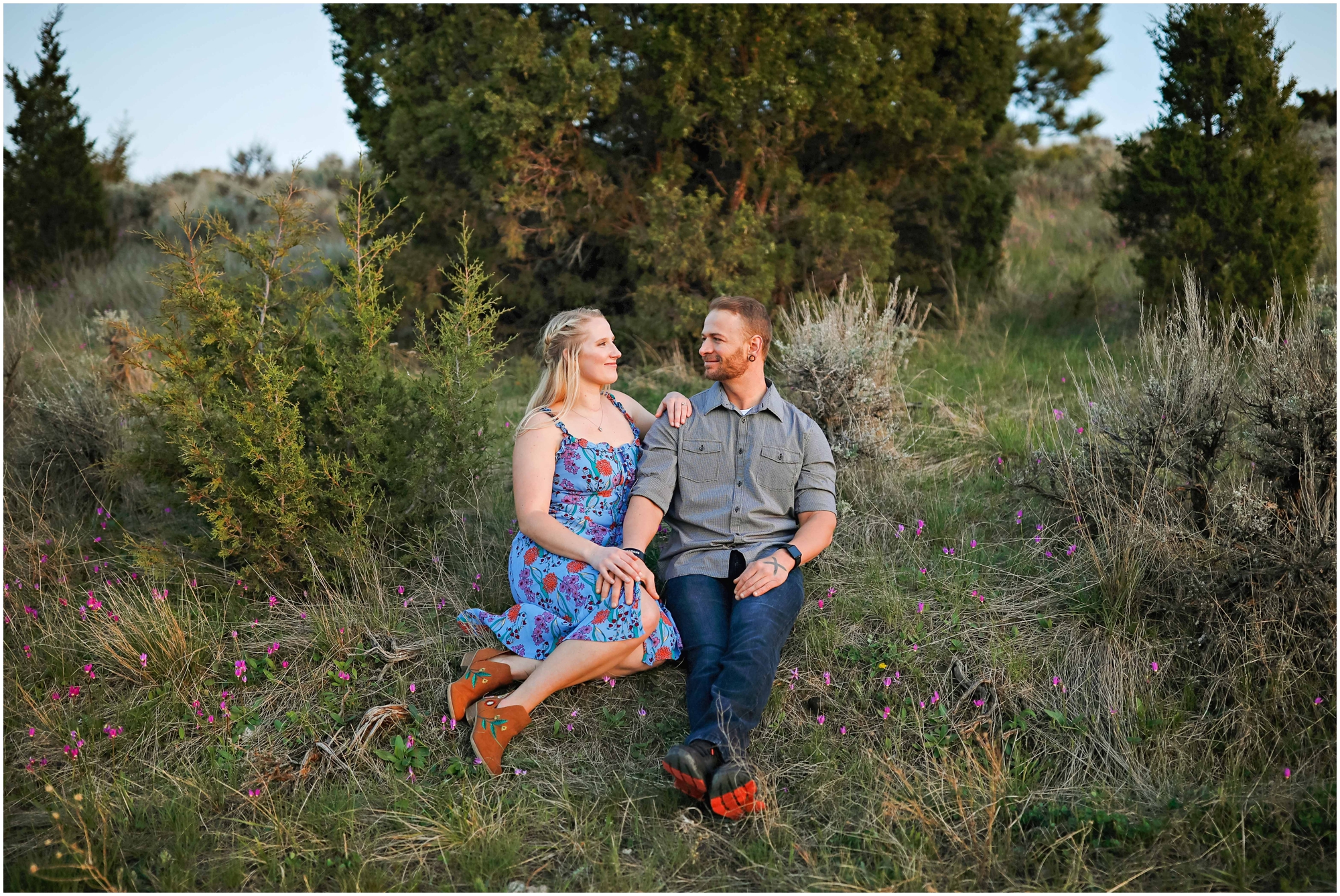 Engaged couple sitting in grassy field with trees and flowers